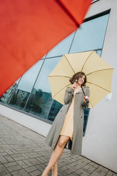都会の背景に黄色い傘で微笑むスタイリッシュな若い女性 — ストック写真