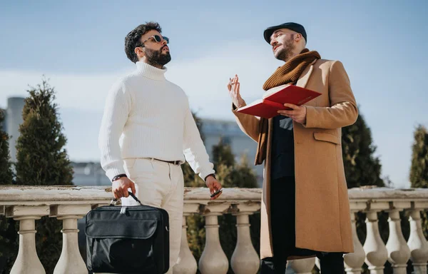 A casual business meeting outside with two fashionable men. One is holding a red notebook and gesturing as he explains something.