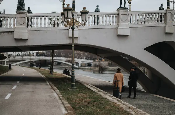 An elegant urban scene capturing two people in deep conversation on a tranquil riverside path with a decorative bridge and lampposts.