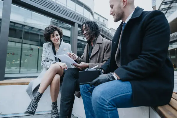 Three diverse professionals engaged in a discussion over paperwork outside of an office building, portraying teamwork and collaboration.