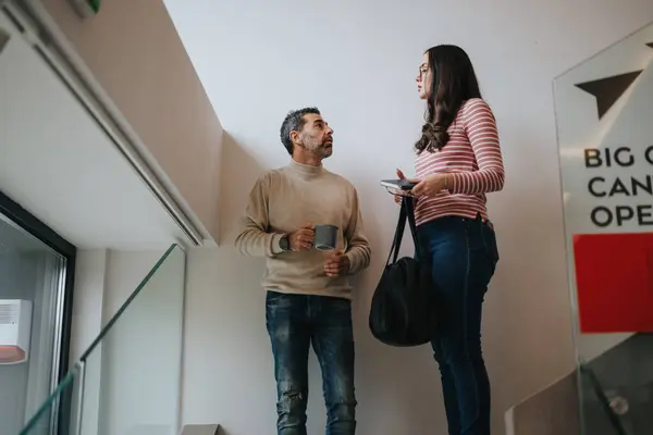 Two professionals engaging in a friendly conversation in an office environment. Man holding a coffee mug with a woman carrying a bag and tablet.