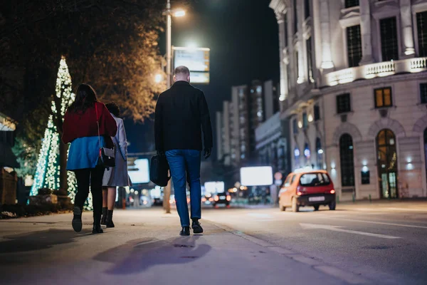 A couple enjoys a nighttime stroll in a lively city, with vibrant Christmas lights adorning the background, adding a festive ambiance to their outing.