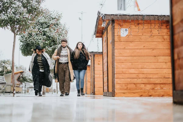 Group of young people in winter clothing walking outside near wooden cabin during a gentle snowfall, embodying teamwork and cold weather.