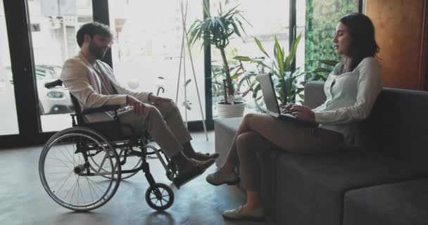 ソファーで休んでいる間 彼女のラップトップに取り組んでいる美しい若い女性と車椅子に座っている障害のある男性の同僚と話し合う — ストック動画