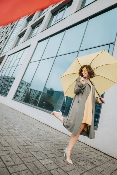 明るい黄色の傘を持つ楽しい女性は 現代の街並みの中でポジティブでスタイルを誇る都市の通りで微笑んでいます — ストック写真