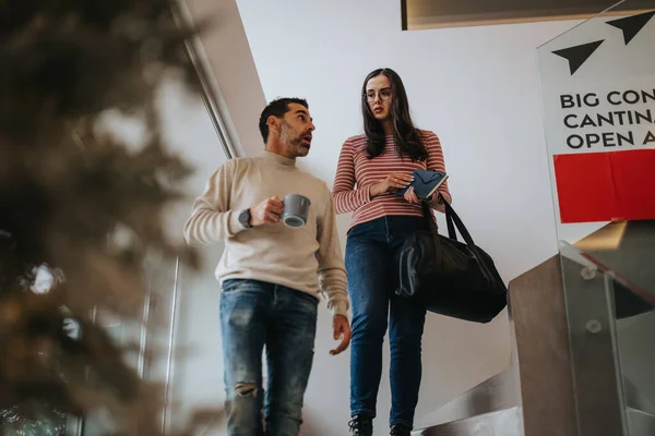 Two business professionals in casual attire casually converse while walking down the stairs in an office building, cups in hand.