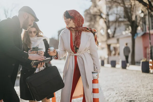 Three friends stand together on a city street, sharing and viewing content on a smart phone. The sunlight filters through, illuminating their casual street wear.