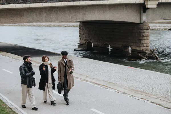 Three fashionable friends engaged in conversation while taking a leisurely stroll by a river, with an old bridge in the backdrop, exuding a sense of calm and camaraderie.