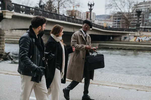 Three diverse professional colleagues engage in a conversation while walking along a riverbank in cold weather, showcasing teamwork and city life.