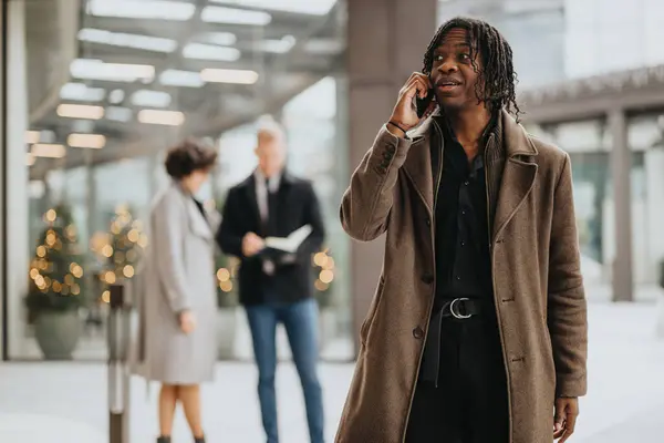 A stylish man engaged in a phone conversation outside, with colleagues and Christmas decorations blurred in the background.