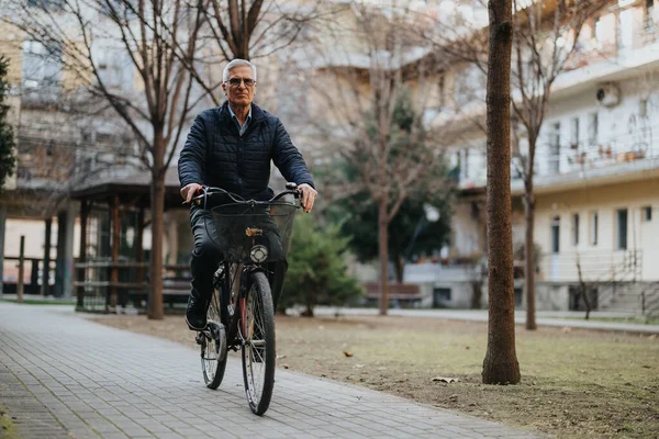 Active older man cycling along a serene park path with trees and apartment buildings in the background, depicting healthy aging and lifestyle.