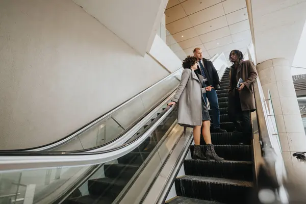 三个穿冬衣的多种族商人在同时代的办公大楼内的自动扶梯上讨论 表现出团队精神和合作精神 — 图库照片
