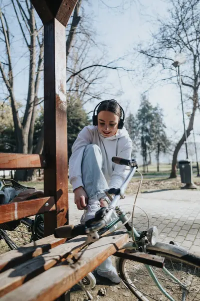 一位年轻女子在公园里享受着阳光灿烂的一天 准备骑自行车 她戴着耳机 把鞋带绑在自行车旁边的长椅上 — 图库照片