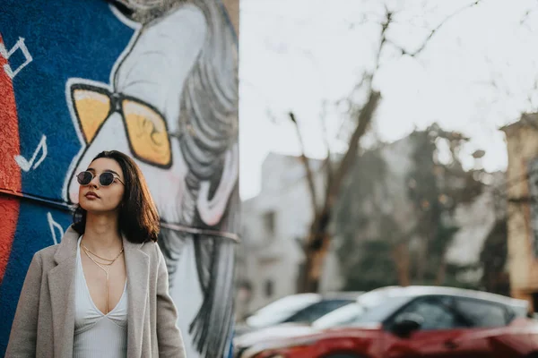 A fashionable young girl walks through the city streets, showcasing autumn vibes with her stylish outfit and confident stance against a backdrop of urban art.