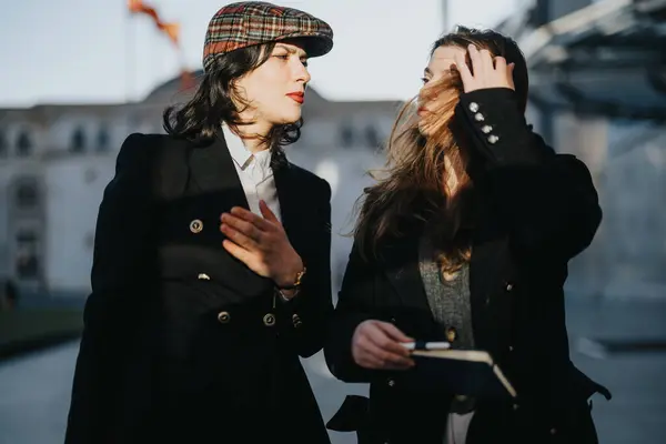 Elegant young women with a notebook walking and talking on a sunny winter day in an urban setting, displaying friendship and style.