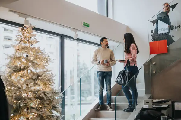 Two engage in a casual conversation on an office staircase during the holidays, with a festive Christmas tree adorning the scene.