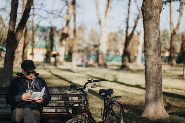 一位年长的绅士在公园里的一条阳光普照的长椅上放着一本书放松下来 他的可靠的自行车站在他身边 描绘着退休后的休闲与宁静 — 图库照片