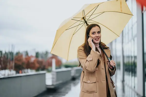 On a cloudy day, a young woman chats happily on her smart phone, holding a sunny yellow umbrella, creating a contrast with the grey backdrop.