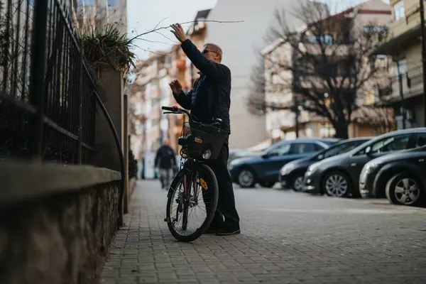 An active senior man in a casual outfit on an urban street, balancing on his bicycle while reaching out for a tree branch.