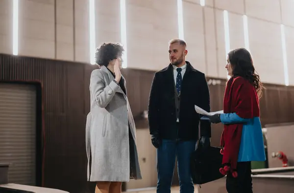 Three business colleagues are engaged in a conversation outside a modern building. They are dressed in warm winter attire, indicating a cold weather meeting.