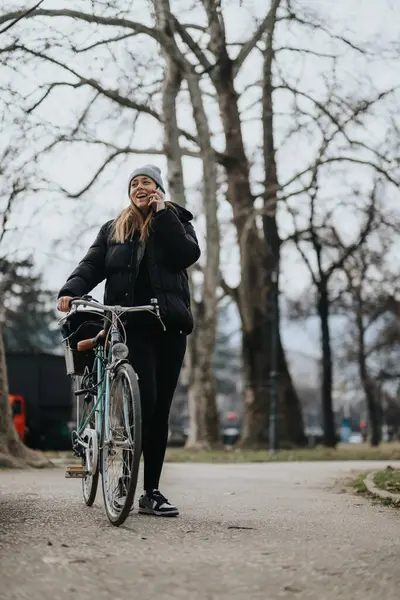 ビーニーとジャケットを着た陽気な若い女性は バックグラウンドの裸の木で公園の道に彼女のヴィンテージバイクと立っている間 電話でチャット — ストック写真