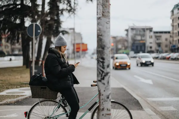 一位身着冬装的年轻成年女子在路边停下脚步 一边骑着自行车 一边在城市里与智能手机交流 — 图库照片