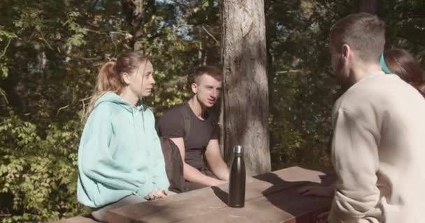 若いスポーツ選手は 自然の中でハイキングや運動から休憩を取る山岳公園のテーブルに集まりました 彼らは座って会話をする — ストック動画