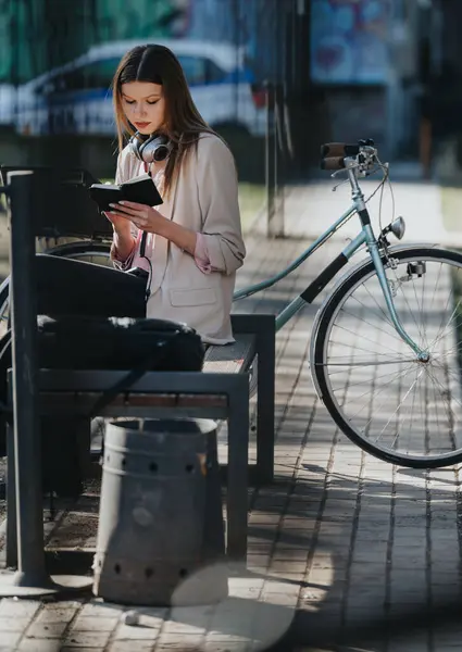首と自転車の周りのヘッドフォンでバスの停留所で読書を集中させた若い女性は 都市の設定で近くに駐車しました — ストック写真