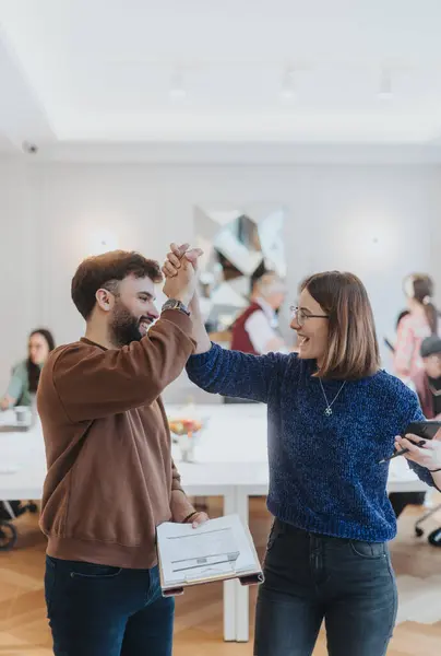 Joyful professional interaction captured as two colleagues high-five in a well-lit office setting, symbolizing team success, support, and positive workplace culture, with other people in the