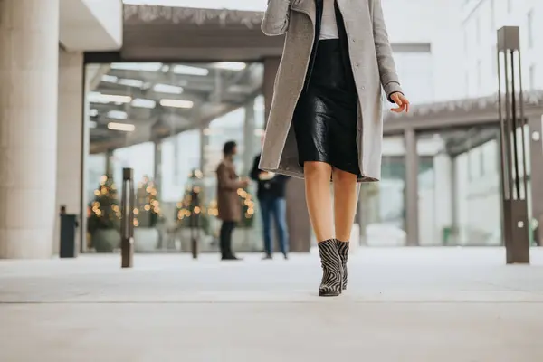 A chic woman in a leather skirt and ankle boots strides confidently through an urban building entry adorned with twinkling Christmas lights.