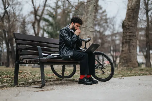 一位穿着时髦服装 专心致志的商人坐在公园的长椅上 坐在膝上型电脑上远程办公 他的自行车就在旁边 — 图库照片