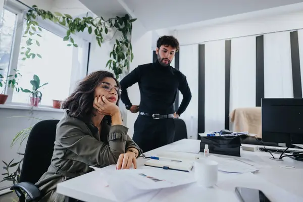 彼女の机で考えている高価な女性 男性の同僚が明るい現代的なオフィス環境で近くに立っている間 — ストック写真