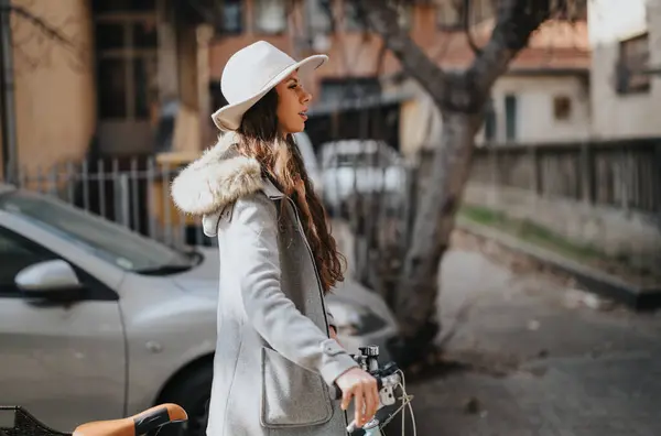 穿着时髦外套 头戴帽子的优雅的年轻女子骑着自行车走在城市街道上 体现了一种休闲而现代的生活方式 — 图库照片
