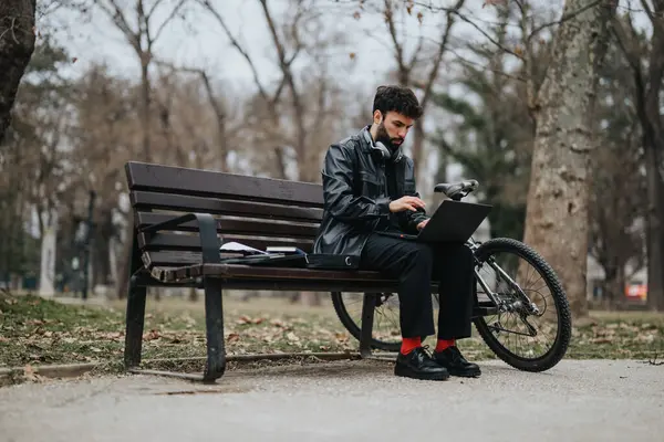 专注的男性企业家使用笔记本电脑坐在公园的长椅上 身边有自行车 体现了专业精神和休闲户外工作环境的混合 — 图库照片