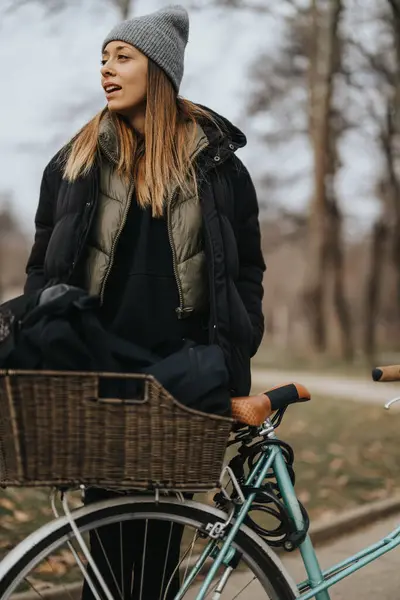 暖かいビーニーとスタイリッシュな女性は 秋のシーズン中に穏やかな公園の設定でウィッカーバスケットで自転車に乗っている間笑顔 — ストック写真