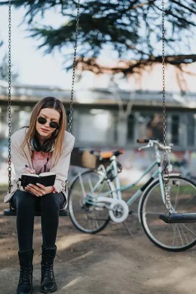 スイングに座りながら読書に吸収されたトレンディな女性を着たカジュアルな屋外レジャーコンセプト 後ろに駐車された自転車 — ストック写真