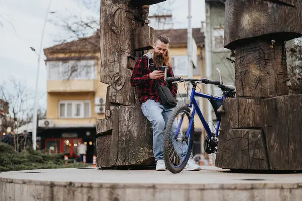 Vousatý Muž Bicyklem Zastaví Městském Prostředí Dívá Telefon Opírá Sochu — Stock fotografie
