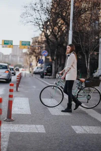 A young, fashionable woman pauses while walking with a classic bike across an urban street setting.