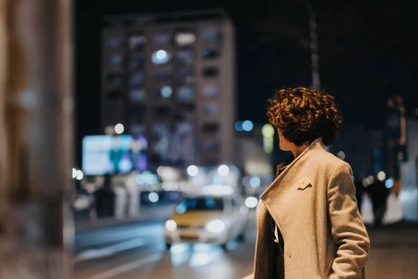 Side profile of a stylish woman in a coat looking at a citys bustling nightlife. The blurred background shows a vibrant urban atmosphere.