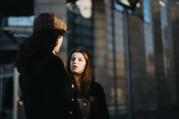 Two young women engaged in a conversation on a city street, with warm sunlight casting shadows around them, portraying a sense of casual urban life.