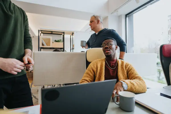 专注的戴眼镜的非洲男人在笔记本电脑上工作 而同事们则在明亮的办公室背景下讨论 描绘团队合作和企业生活方式 — 图库照片