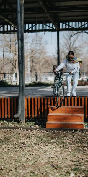 Outdoor shot of a focused young woman carefully carrying her bicycle down a set of wooden stairs in a sunlit urban park.