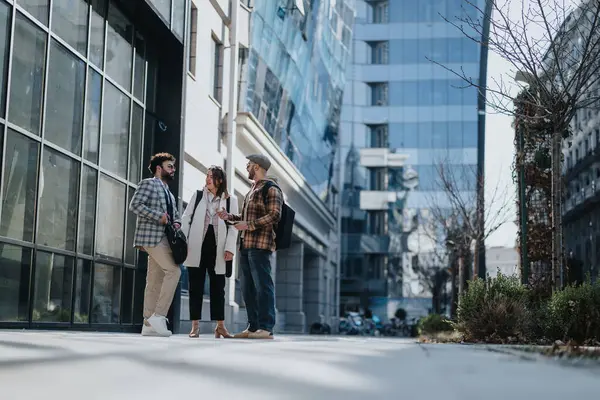 三位年轻的商业专家站在一座现代化的大楼外 就一个项目进行协作 并讨论市场趋势 — 图库照片