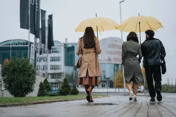 Трое Друзей Идут Дождём Жёлтыми Зонтиками Возле Современных Зданий — стоковое фото