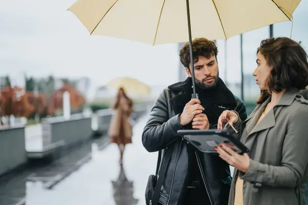 在阴云密布的雨天里 一个男人和一个女人在外面交谈 他们在遮阳伞下审视着一块数码平板电脑 记录了一段团队合作的时光 — 图库照片