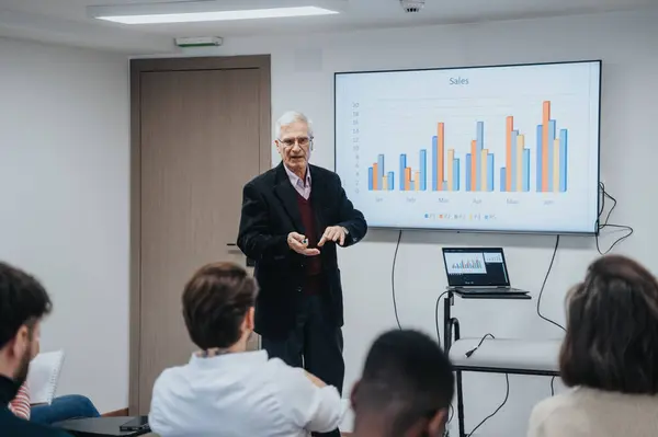 An experienced businessman conducts a sales performance review with a bar chart on the screen in a conference room setting, engaging with attentive listeners.