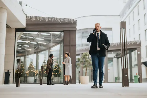 Confident businessman talking on phone walking in urban setting with decorated trees.