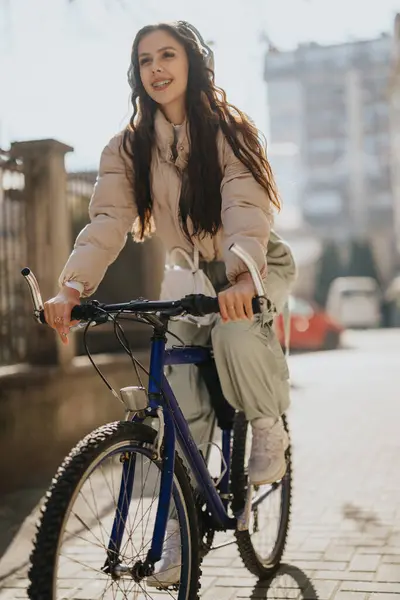 年轻的女骑手带着耳机在城市街道上骑自行车 表达喜悦和积极的生活方式 — 图库照片