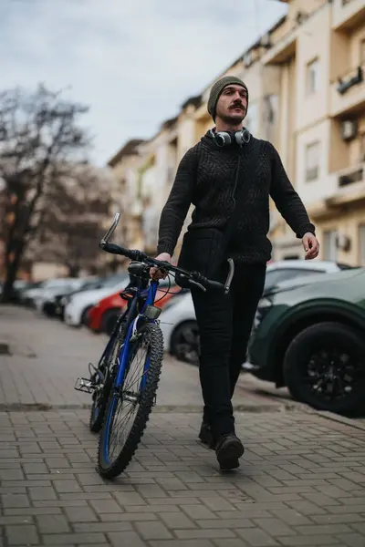 时尚的年轻人 戴着便帽和耳机 骑着自行车在城市街道上散步 流露出一种悠闲的都市气息 — 图库照片