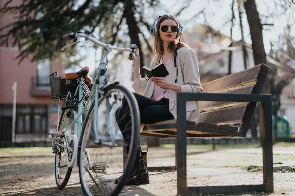 A fashionable woman listens to music with headphones while reading a book on a sunny day in the park, with a vintage bicycle in the foreground.